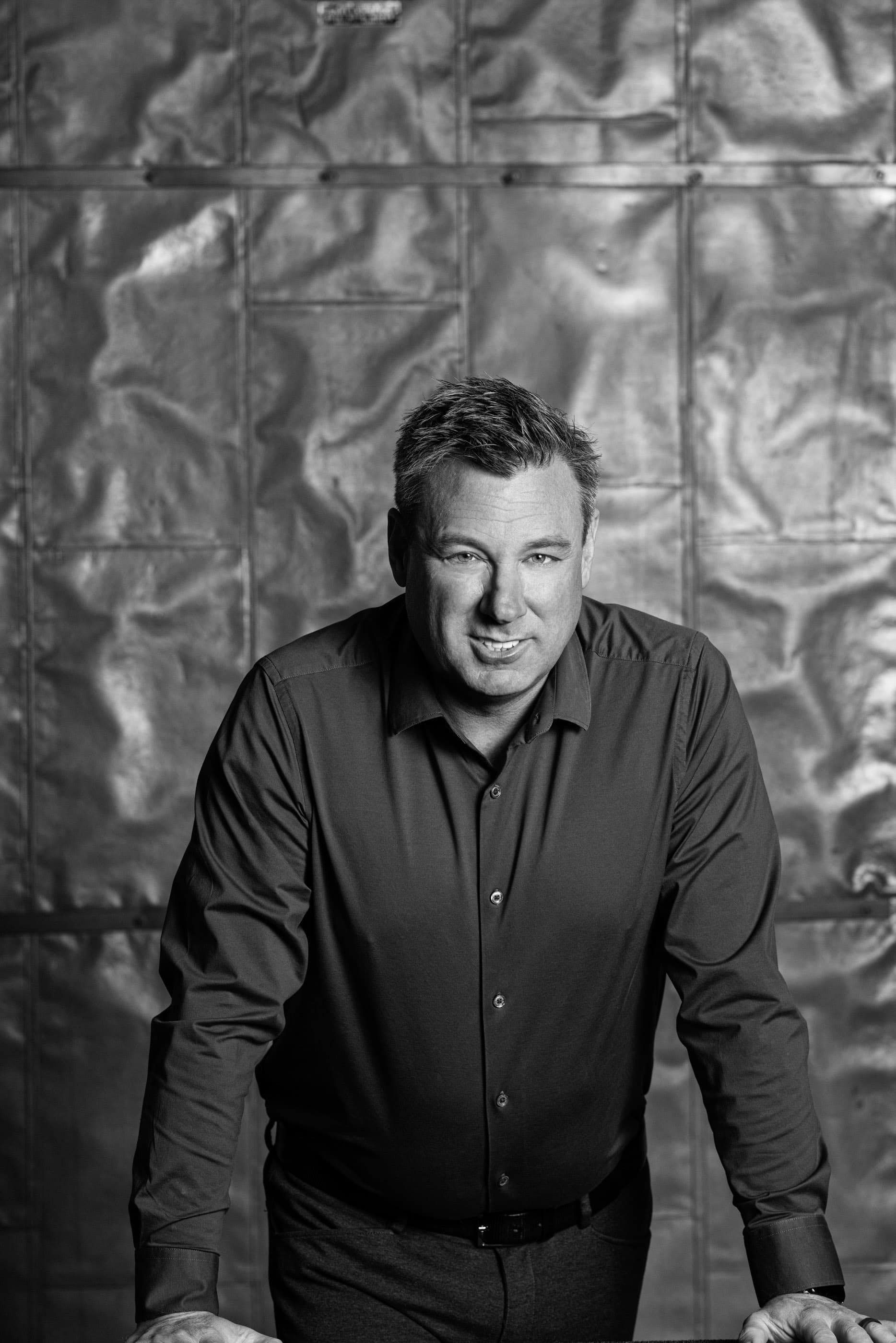 Smiling man in a button-down shirt leaning forward against a textured, metallic paneled wall.