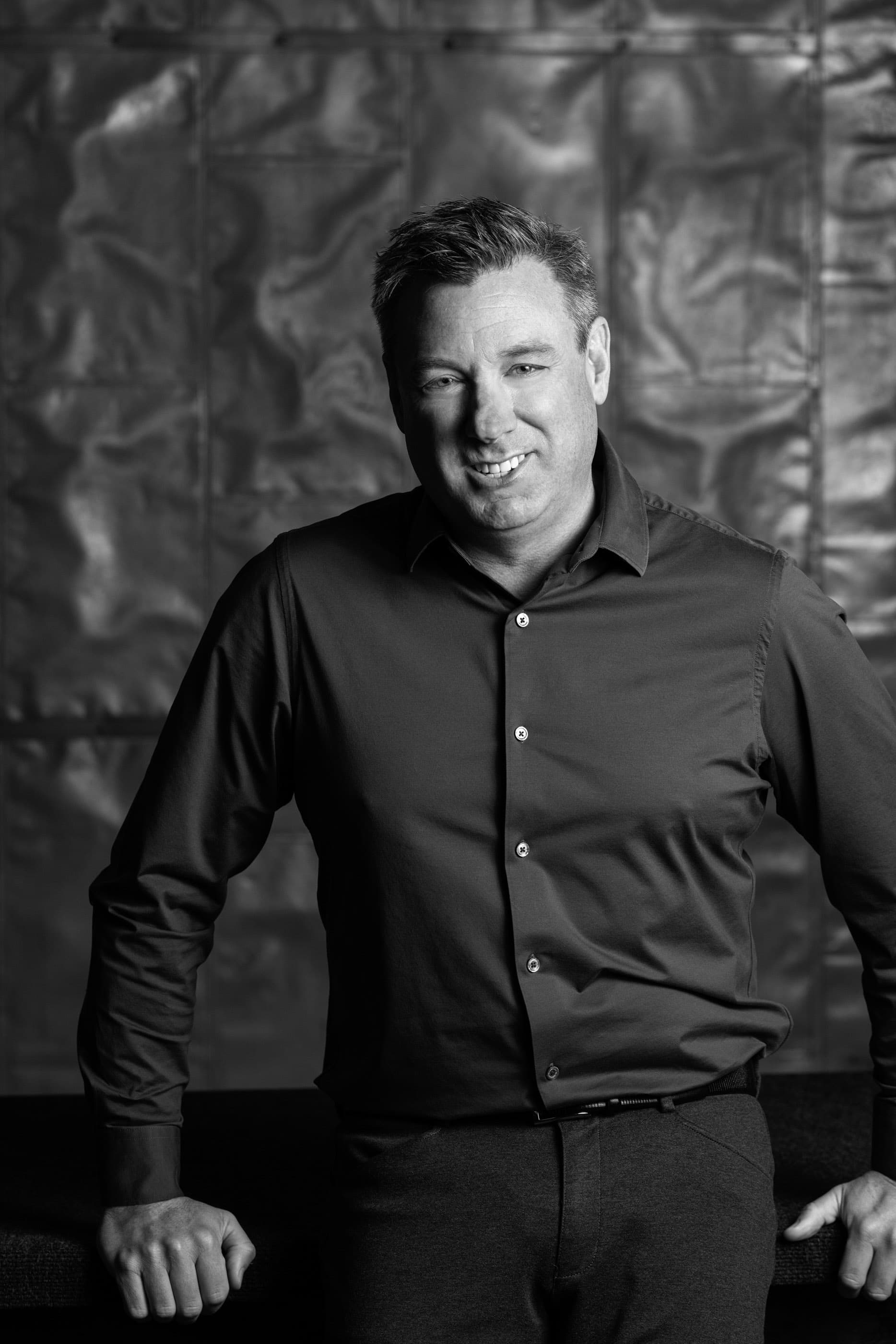 Smiling man in a button-down shirt against a textured metallic wall, black and white.