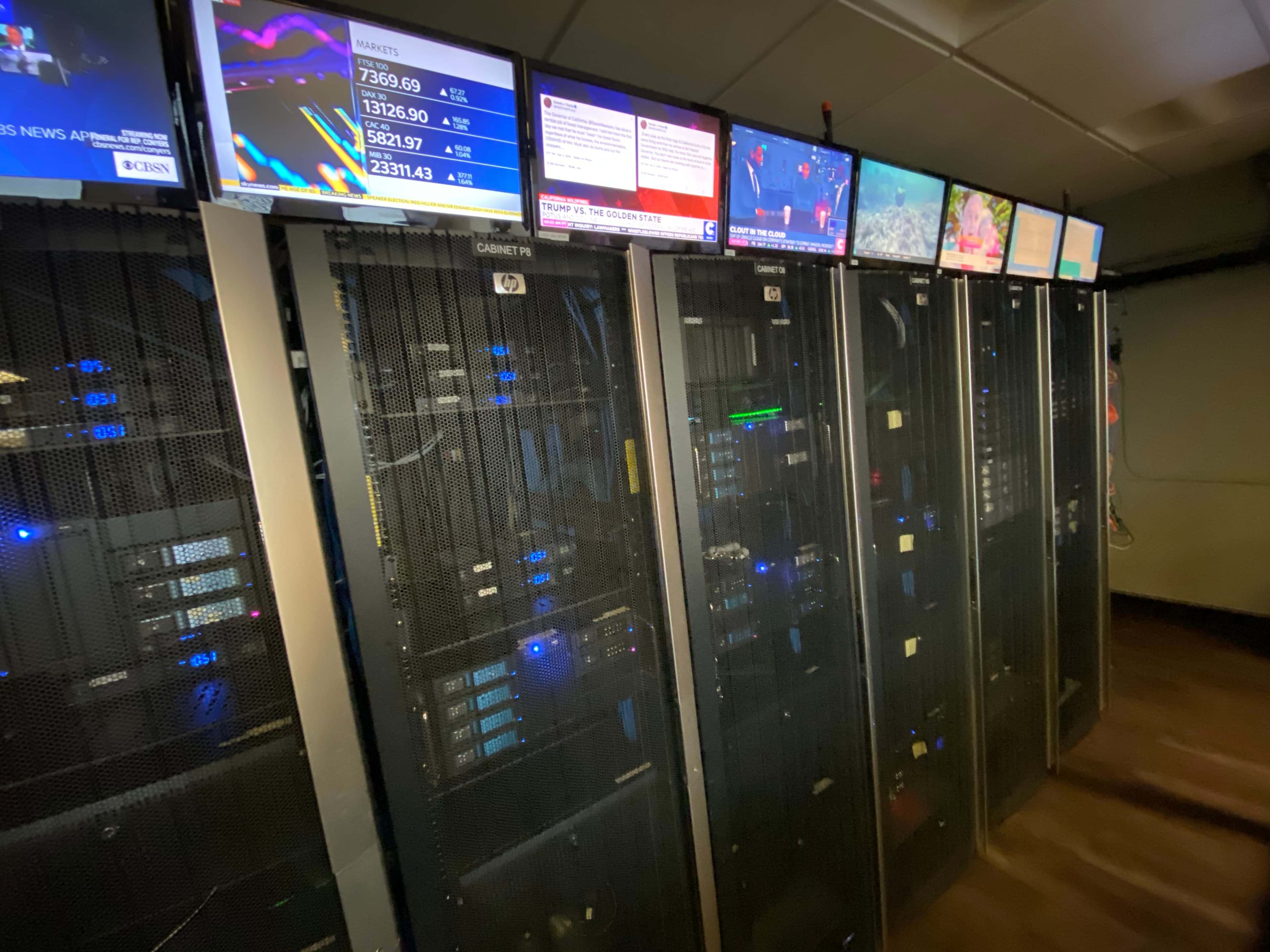 Server racks with blue lights and overhead monitors displaying news and financial market data.