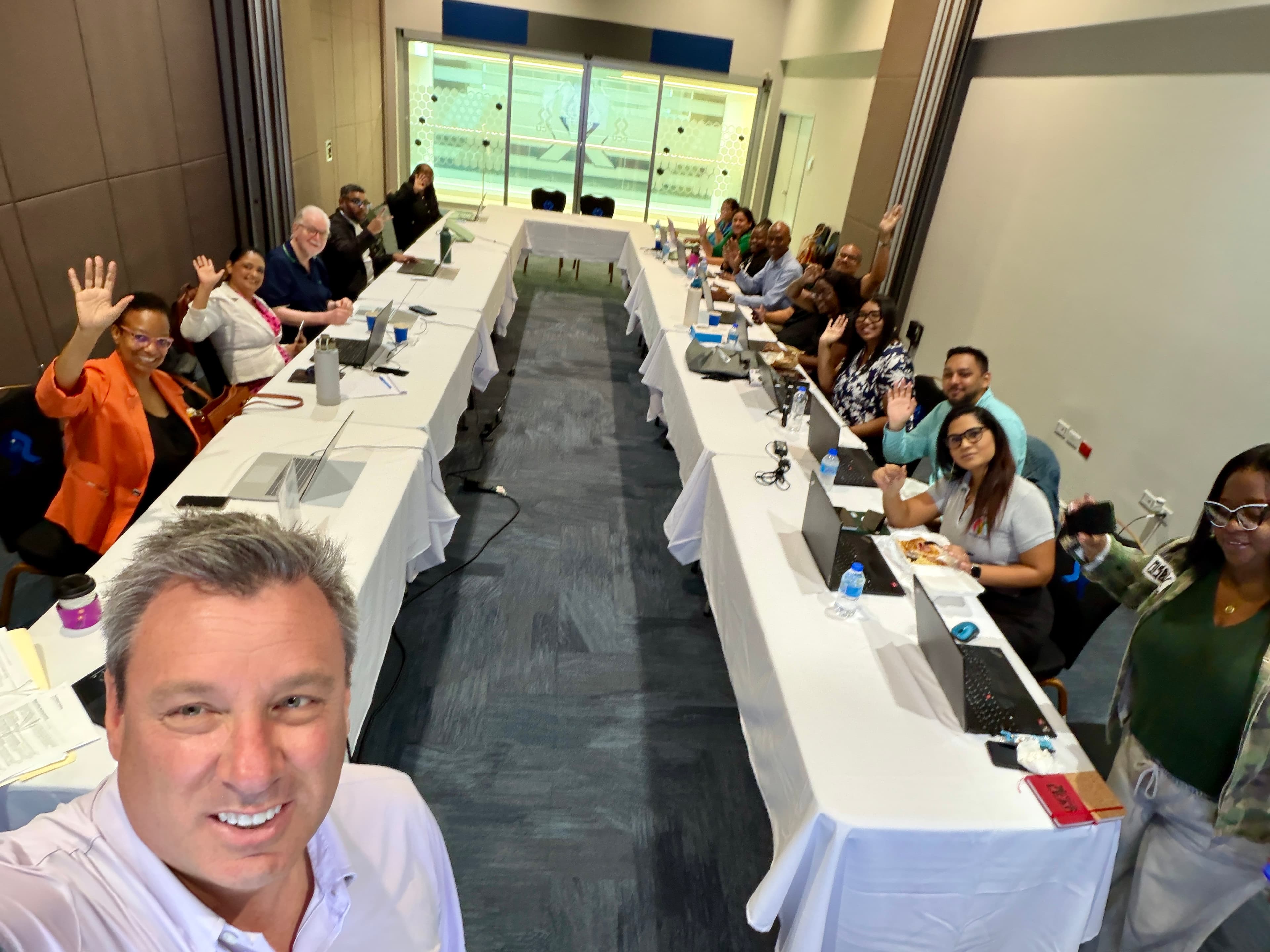 Diverse group of people waving from a U-shaped conference table during a meeting selfie.