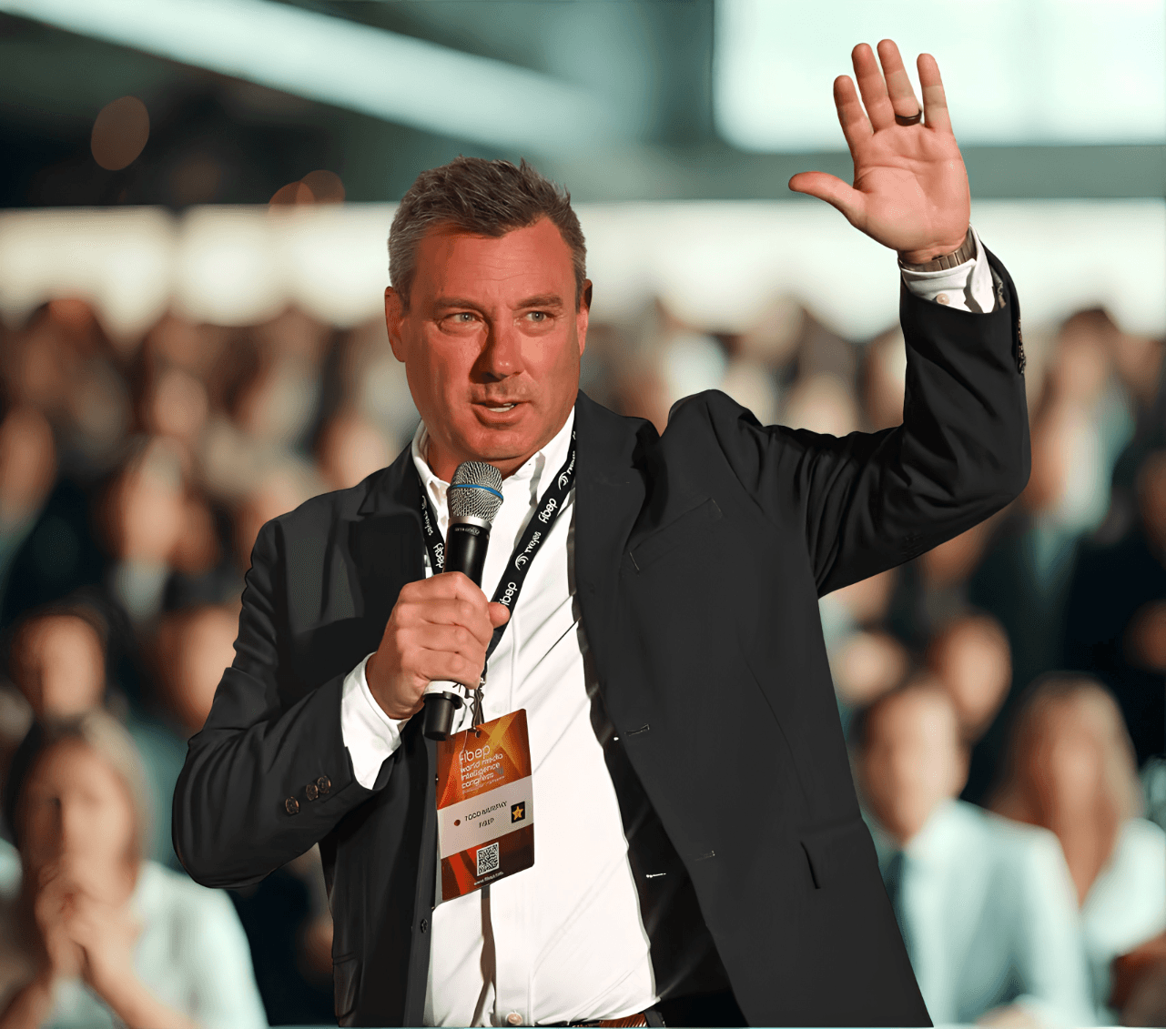 Man in a blazer speaking into a microphone and waving to a large audience.
