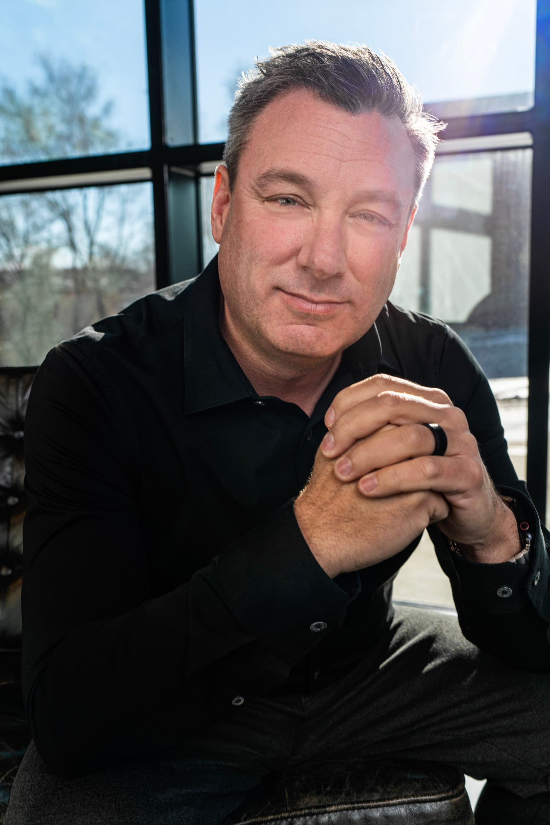 Middle-aged man in a black shirt sits with hands clasped before a sunlit window.