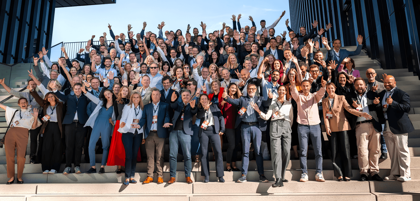 A large, diverse group of smiling professionals waving while standing on wide outdoor stairs.